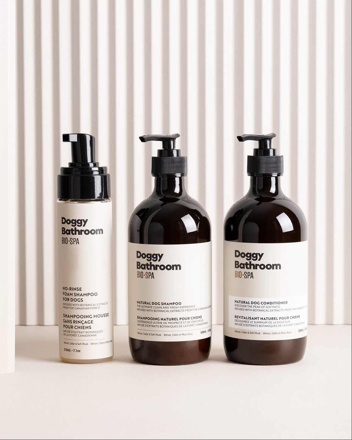 Three Doggy Bathroom pet care bottles arranged on a white surface against a white slatted background: a beige shampoo bottle, a dark amber body shampoo bottle, and a dark amber body conditioner bottle, all with black pump tops and white labels.